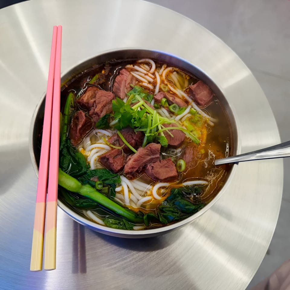 Loong Noodles' beef noodle soup with thick noodles, greens, and pink chopsticks on a metal table.