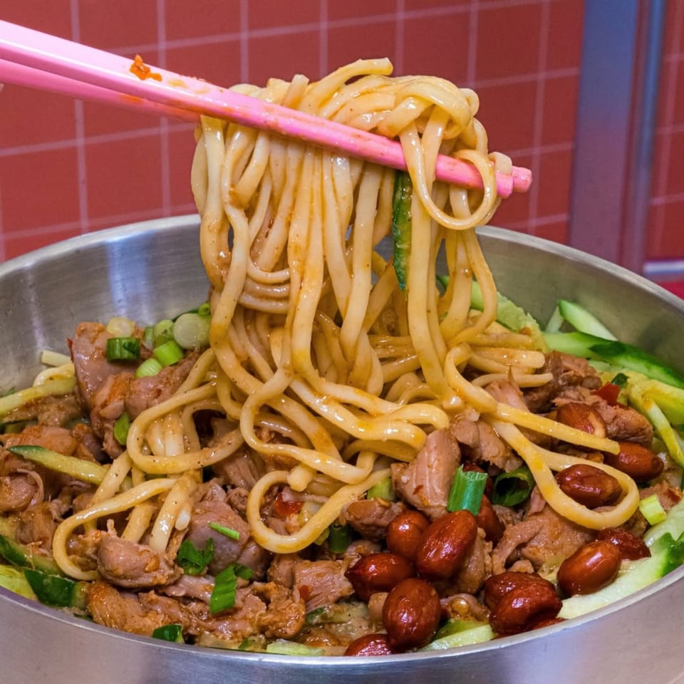 Pink chopsticks lifting saucy Loong Noodles with chicken, peanuts, and cucumber from a metal bowl.