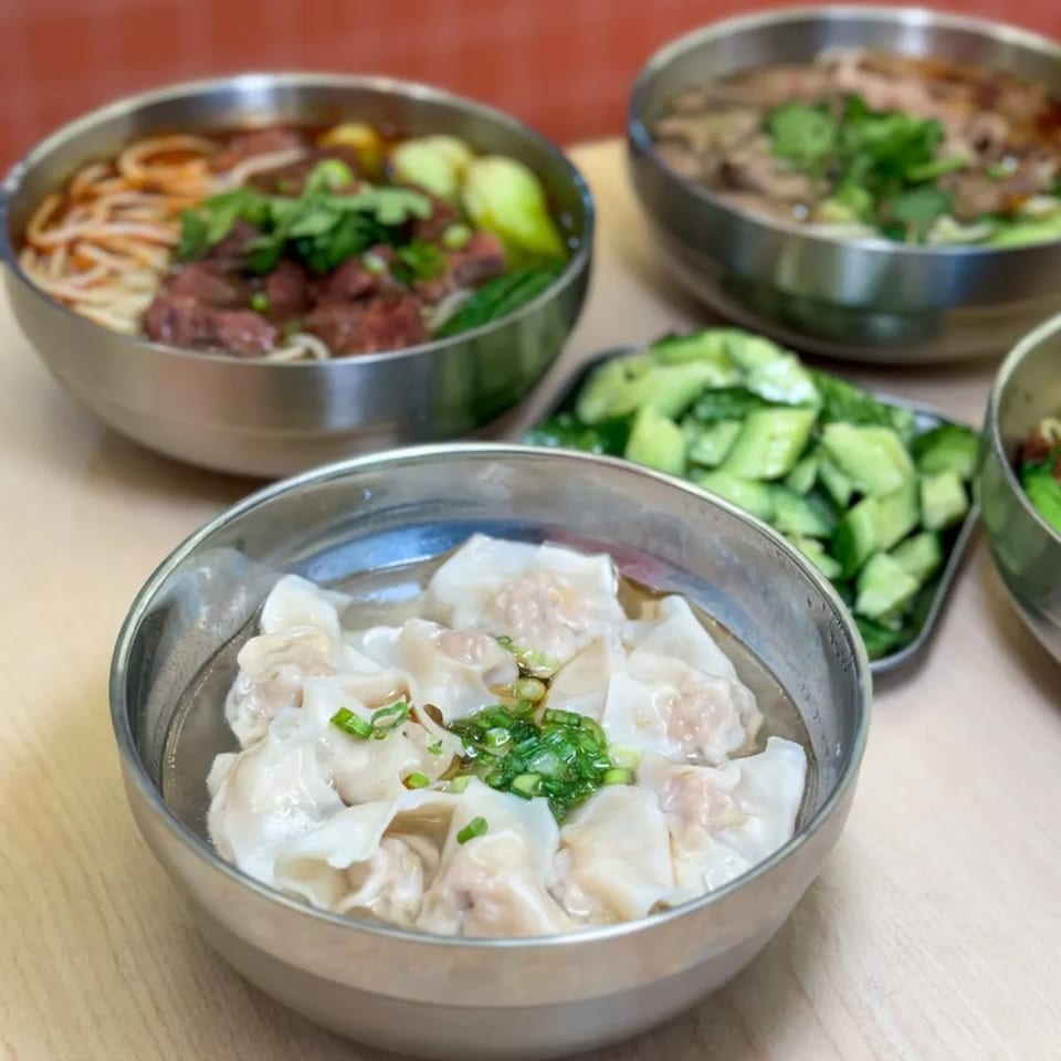 Several bowls of Chinese food, including wonton soup, beef noodle soup, and a side of cucumbers.
