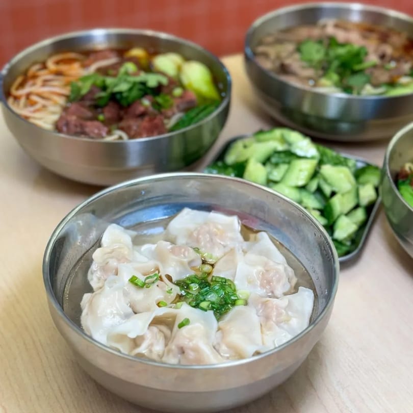 Several bowls of Chinese food, including wonton soup, beef noodle soup, and a side of cucumbers.