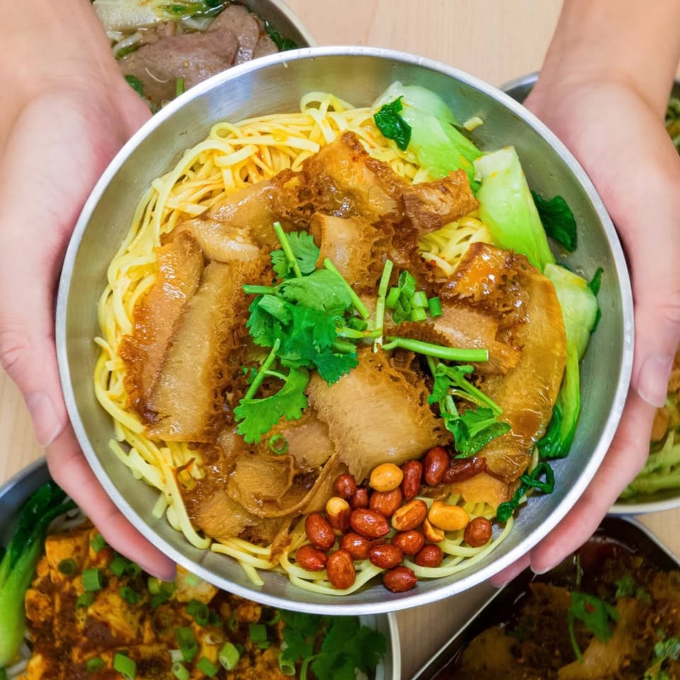 Hands holding a bowl of Loong Noodles with tripe, peanuts, bok choy, and cilantro.